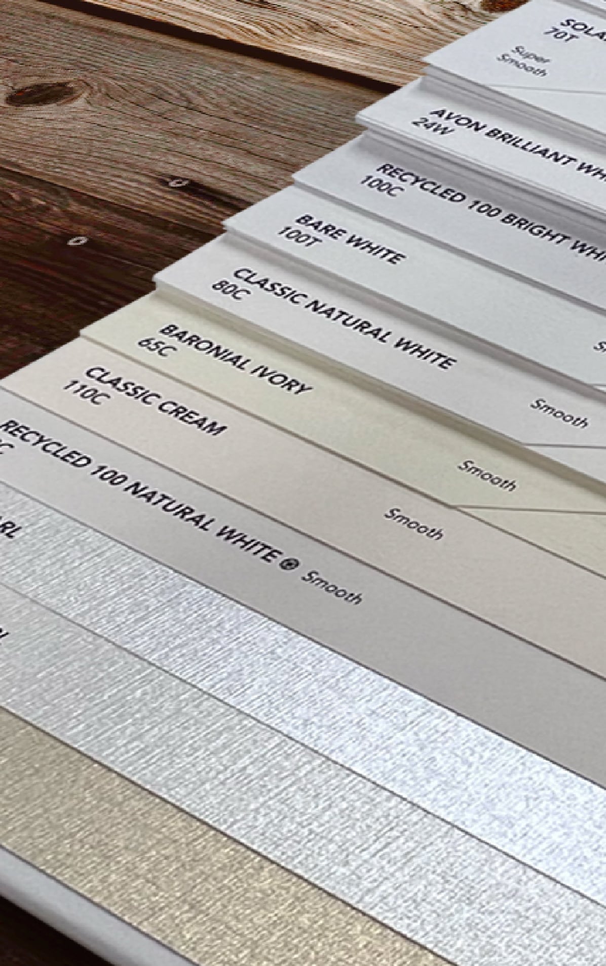 Paper swatch book fanned open on a wood table, each tab labeled with its stock and weight